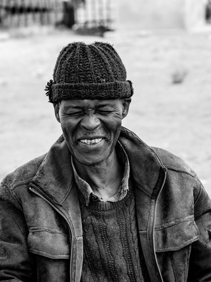 Black and white portrait of a smiling older man in a knitted beanie and leather jacket, from the Optix Legacy series